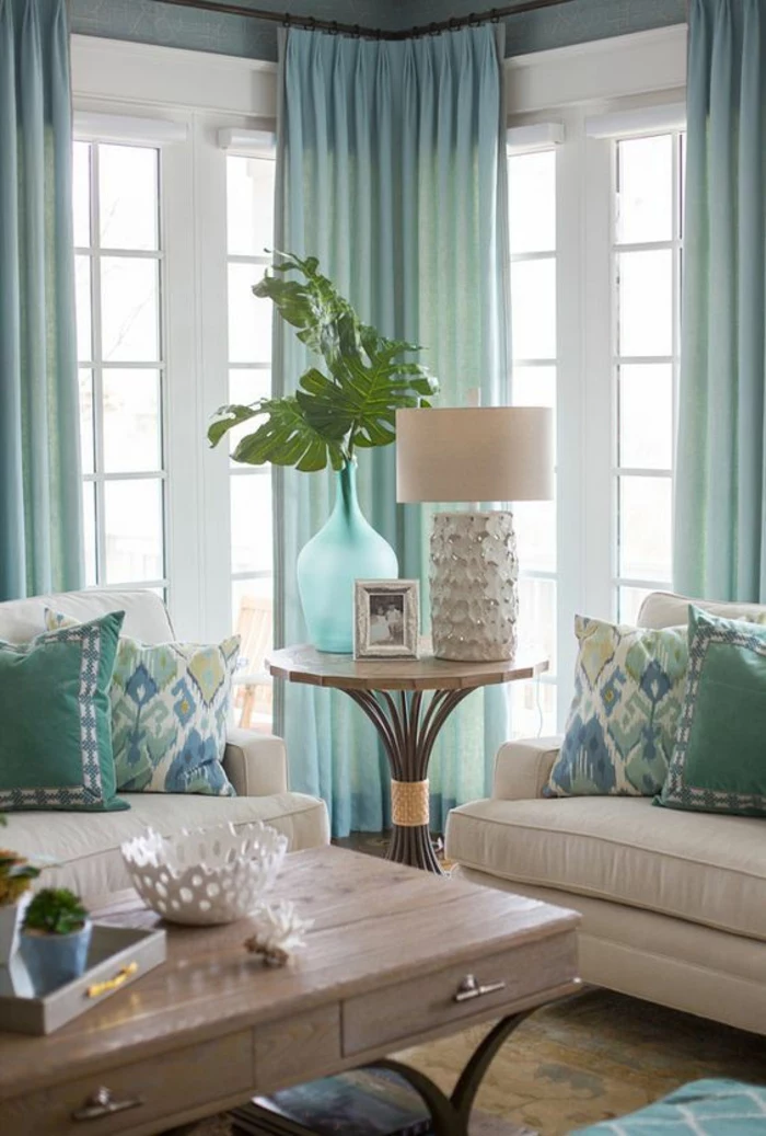 windows with white frames and light blue curtains, two cream colored sofas with blue cushions, light colored wooden table with drawers, round coffee table with blue vase with green plant