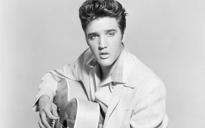 black and white photo of elvis presley, pale blazer and shirt, wavy messy gelled up hair, holding acoustic guitar, playing and singing