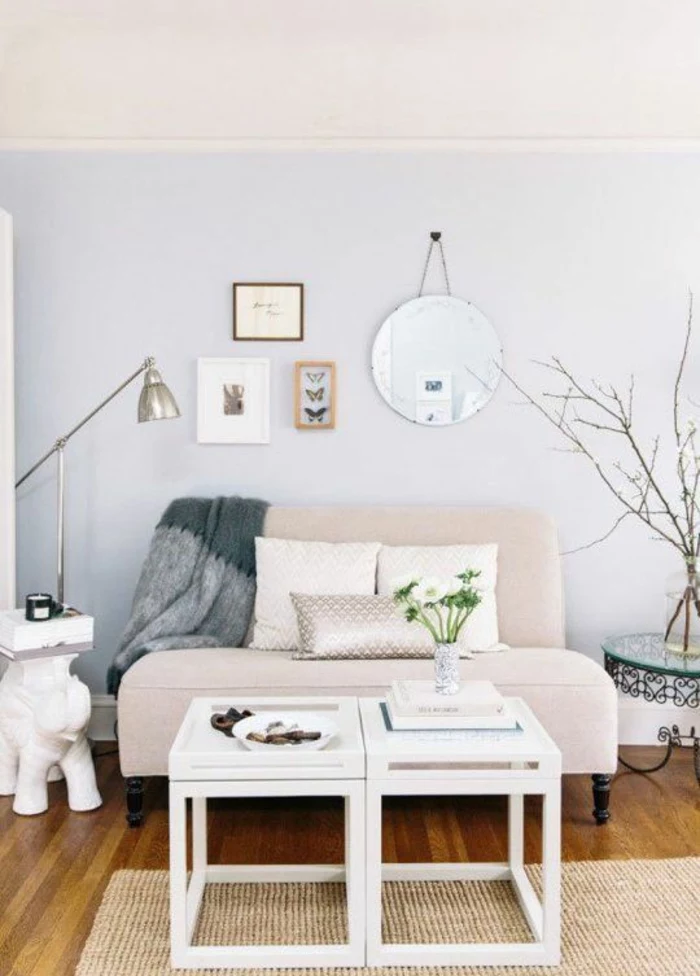 two tone living room walls, pale lilac and cream wall, very pale small pastel pink sofa with three cushions, small white coffee table, wooden floor and cream rug, lamp framed images and grey blanket