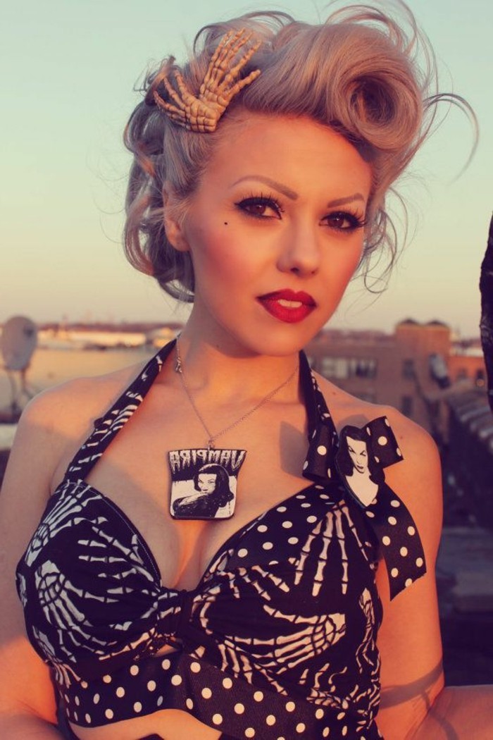 pinup style, goth-influenced platinum blonde woman with black top, white skeleton hands print, vampira necklace and badge, dark red lipstick and black eye make up, victory roll with skeleton ornament