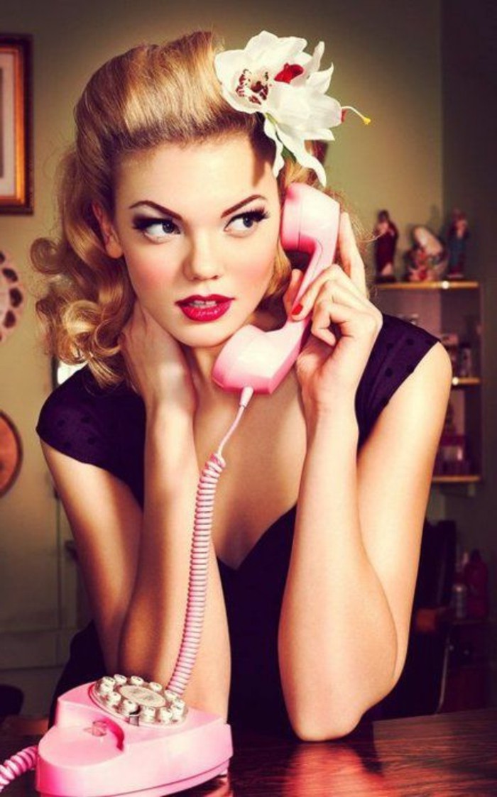 pinup style, blonde woman with fake white flower in hair, heavy make up bright red lipstick, dark black eye makeup with mascara an eyeliner, black top and red nail polish, holding a pink telephone receiver