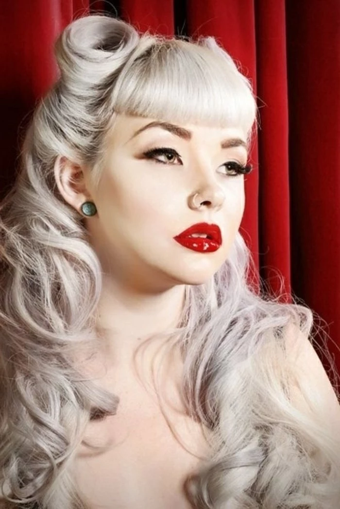 betty bangs, woman with long platinum blonde hair, victory rolls curls and bangs, nude shoulders a nose ring and green stud earrings, red lipstick fake lashes mascara