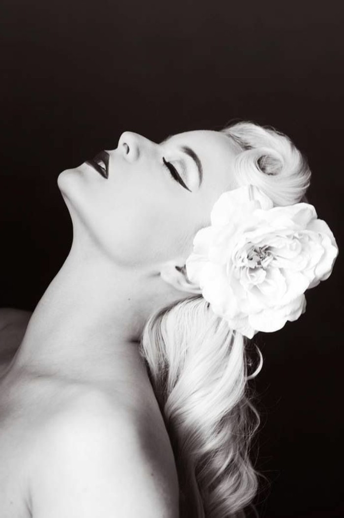 pinup hair, black and white photo of woman in profile facing upwards, light curled hair with big fake white flower, bold eyeliner and lipstick, nude shoulders and black background