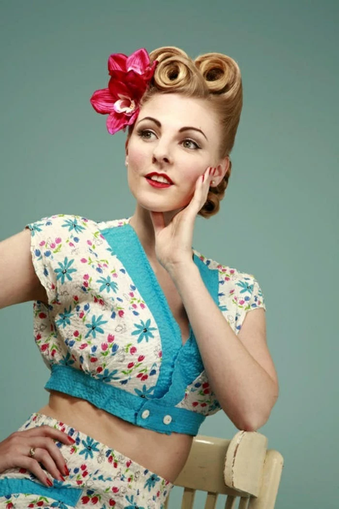 blonde woman leaning on white chair, hair styled in three victory rolls with two fake pink tropical flowers, wearing a two piece suit in white and blue