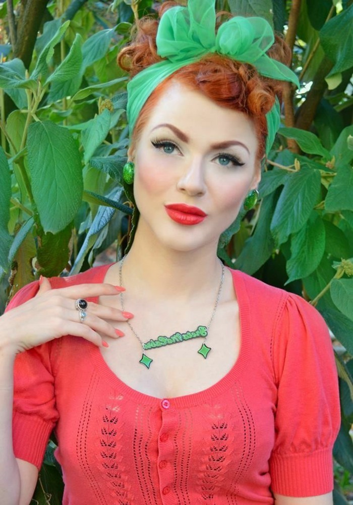 red-haired woman with curly hair, tied up with green scarf, big green earrings and necklace, heavy make up coral pink top and nail polish