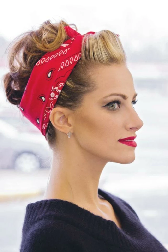 pin up hairstyles, close up of woman in profile, blond hair with curls tied with a red bandanna, red lipstick eyeliner and fake lashes, navy sweater and light background 