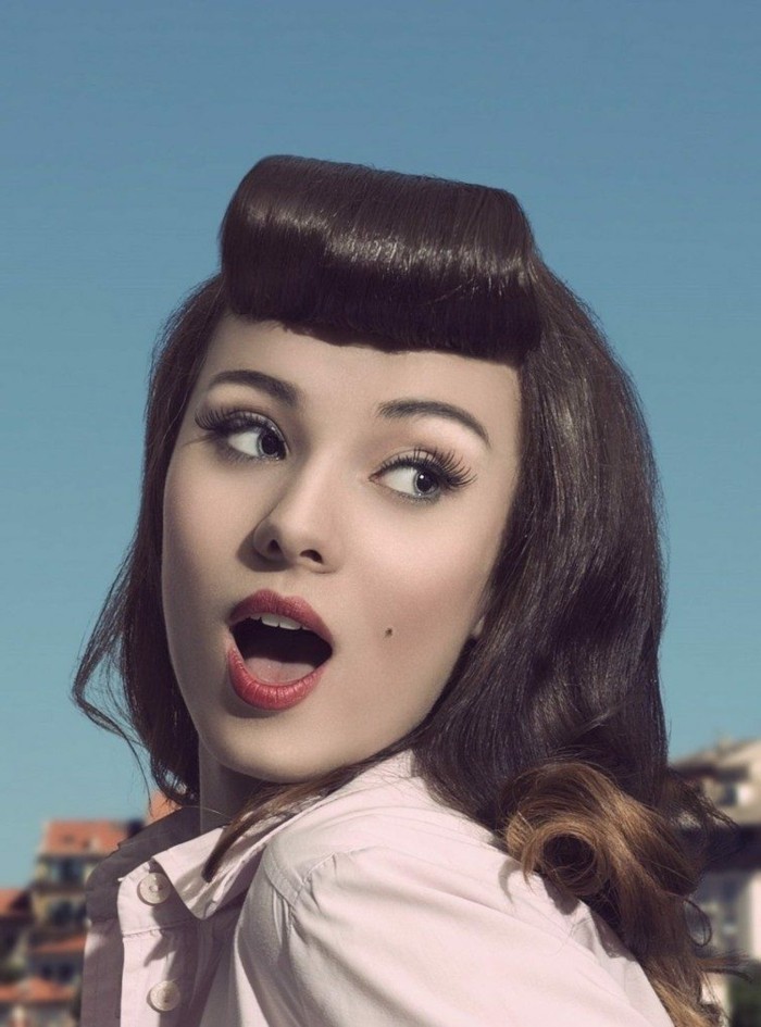 surprised-looking woman in close up, dark hair with retro styled bangs, fake lashes and black eyeliner, red lipstick and a beauty spot, pale pink shirt and blue background