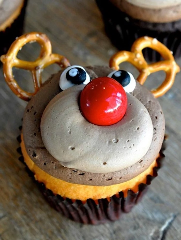 close up of a cupcake with brown icing, made to look like rudolph, with pretzels for antlers, big red candy for nose and frosted eyes