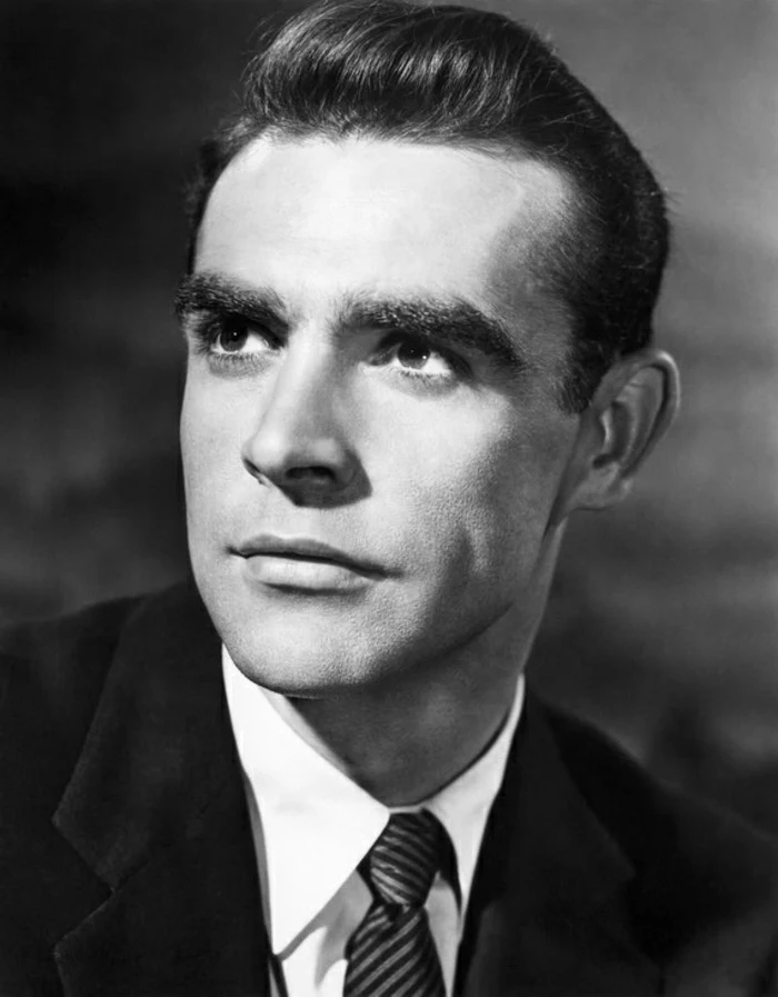 black and white photo of young sean connery, black blazer and white shirt, striped tie and gelled up hair, grey background