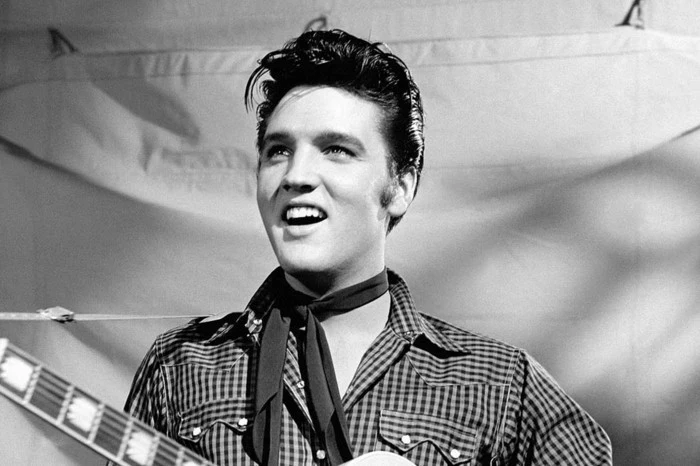 grey scale photo of young elvis presley, gelled up shiny black hair, smiling or singing, chequered shirt with pockets and small narrow black scarf tied around neck