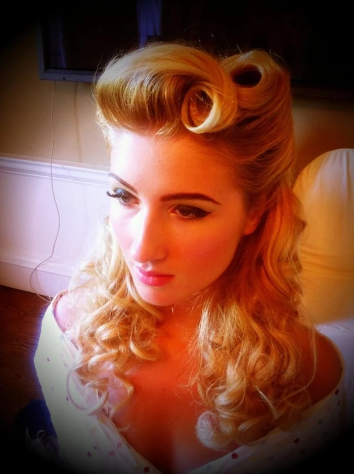 blonde woman seen from above, wearing heavy mascara and fake lashes, curled shoulder-length hair and victory rolls, white top and dark filter