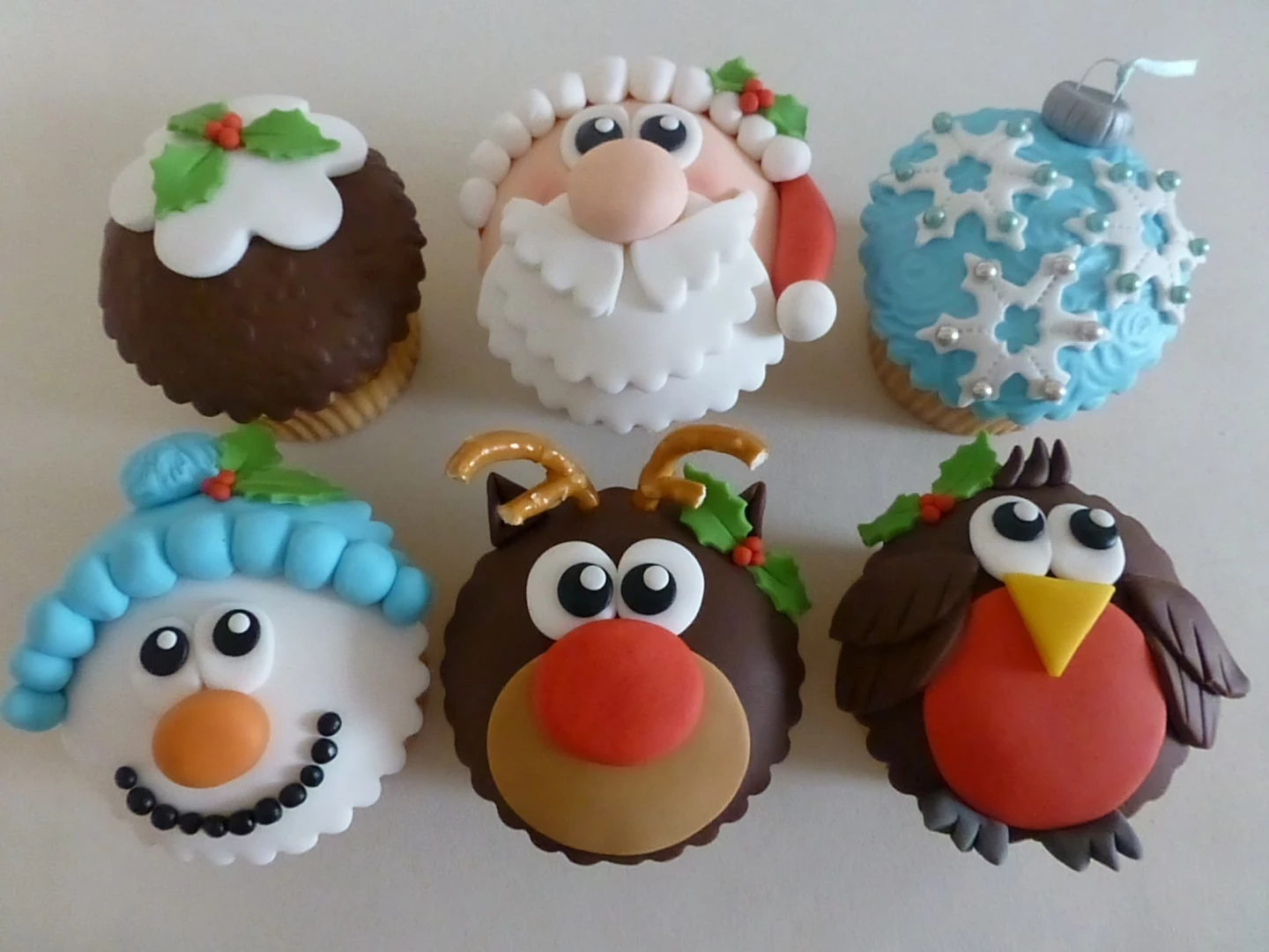 six cupcakes decorated with brown, white red pink and blue fondant icing, shaped like santa and reindeer, snowman and bird, christmas pudding and ornament