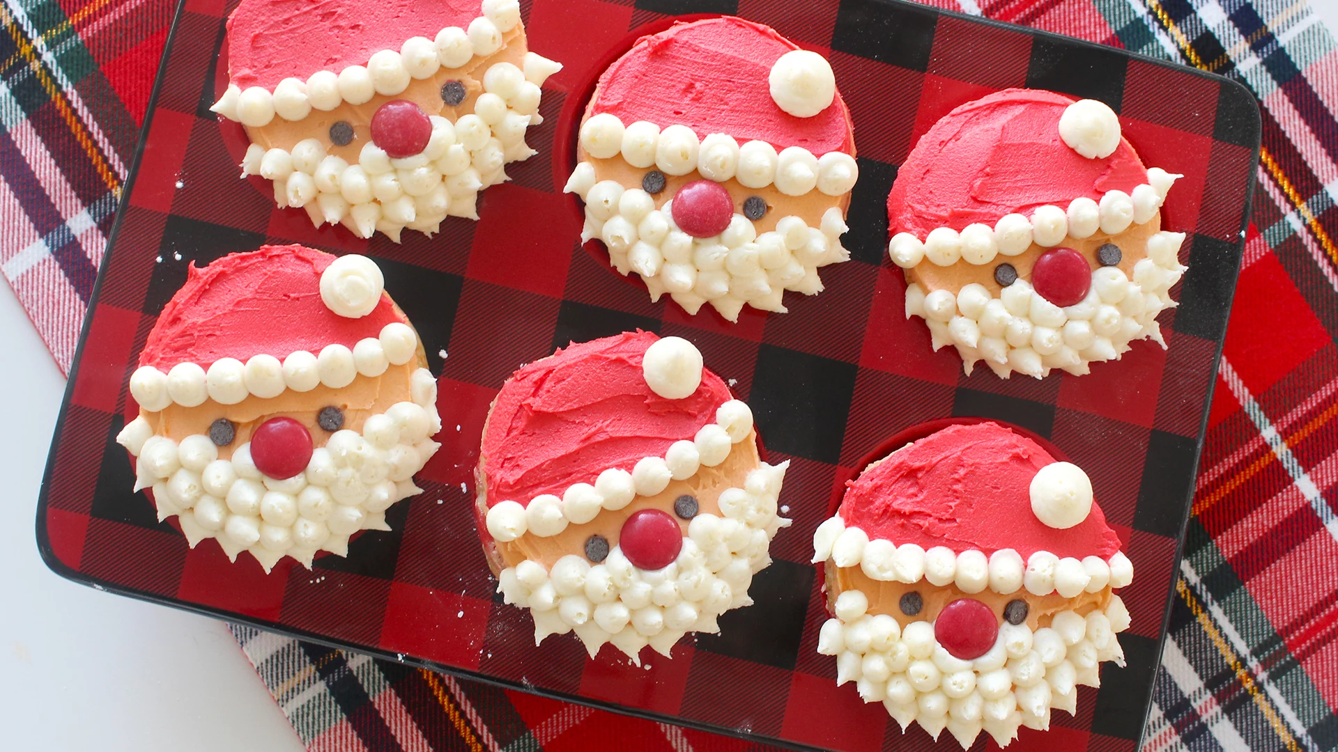 six cupcakes on red and black checkered muffin mould, decorated with red white and orange frosting, made to look like santa, red candies for noses