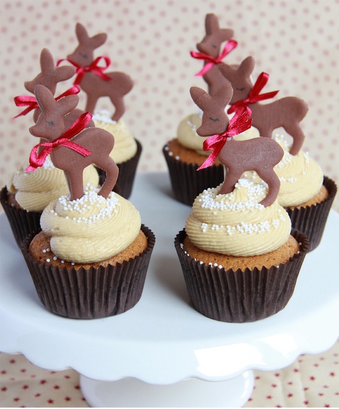 six cupcakes in brown wrappers, with light yellow frosting, decorated with chocolate deer shapes, with red bows around their necks