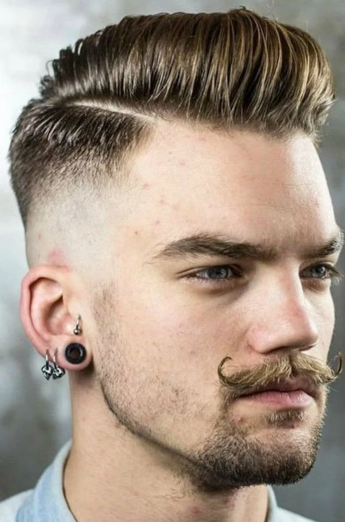 side part undercut, man with fair hair and twisted up mustache, stubble on chin and three earnings, one flesh earring and pale top, hair parted on the side, longer on top but shaven on sides