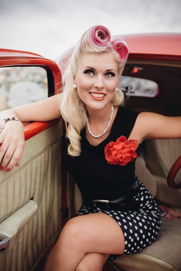 pin up hairstyles, smiling woman sitting in red retro car, black tank top and skirt with white polka dots, faux red flower and pearl necklace, platinum blonde hair in 1950's style with pink dye