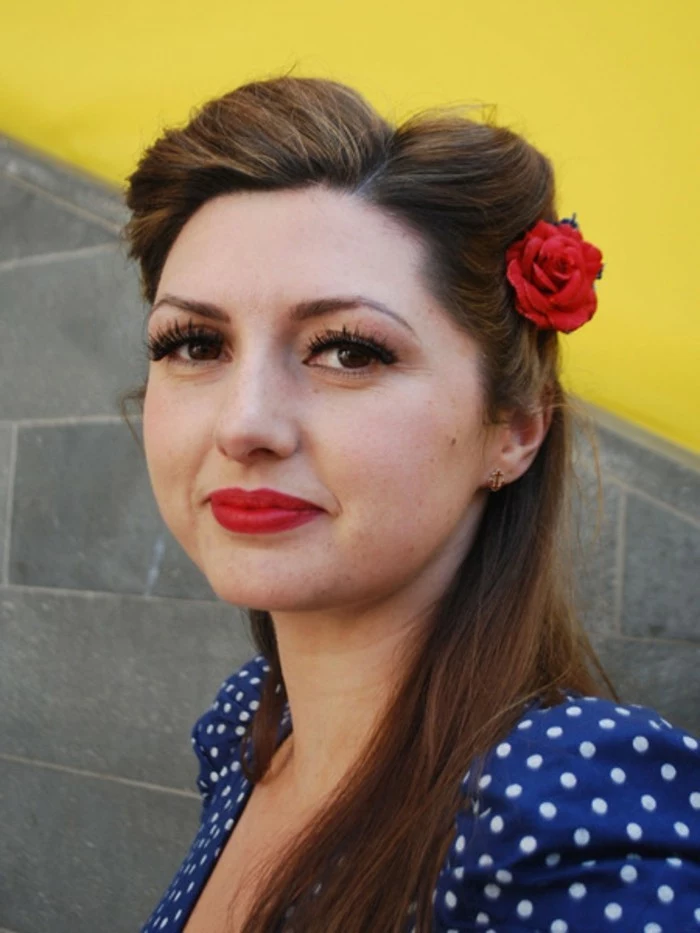 pinned up hairstyles for long hair, woman wearing a blue top with white polka dots, brown hair with retro twist and a fake red flower, fake eyelashes mascara eyeliner and red lipstick