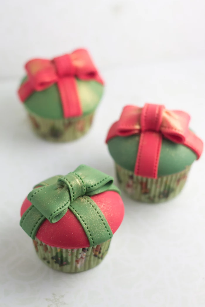 three cupcakes in green festive wrappers, green or red fondant frosting, red or green fondant bows on top, pale background