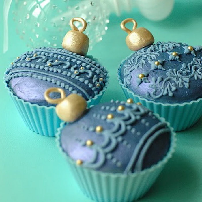 three cupcakes in pale blue moulds, with dark blue and gold icing, made to look like christmas ornaments, light turquoise background