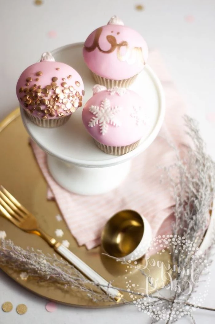 three cupcakes with pink fondant frosting, decorated with golden letters and sprinkles, and white snowflakes, on white dish and golden tray