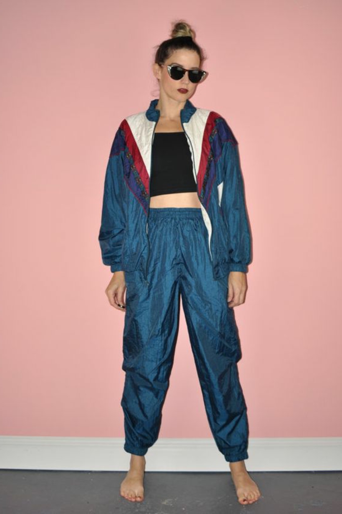 what did people wear in the 80s, woman with top knot and dark retro sunglasses, wearing a shiny windbreaker, track bottoms and black crop-top, barefoot on grey floor with pink background