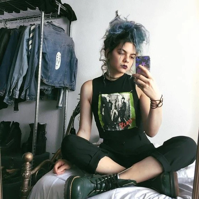 what did people wear in the 80s, young punk girl with died blue hair and dark lipstick, wearing black cropped pants and band t-shirt, with dark green army boots, sitting cross-legged on bed holding phone