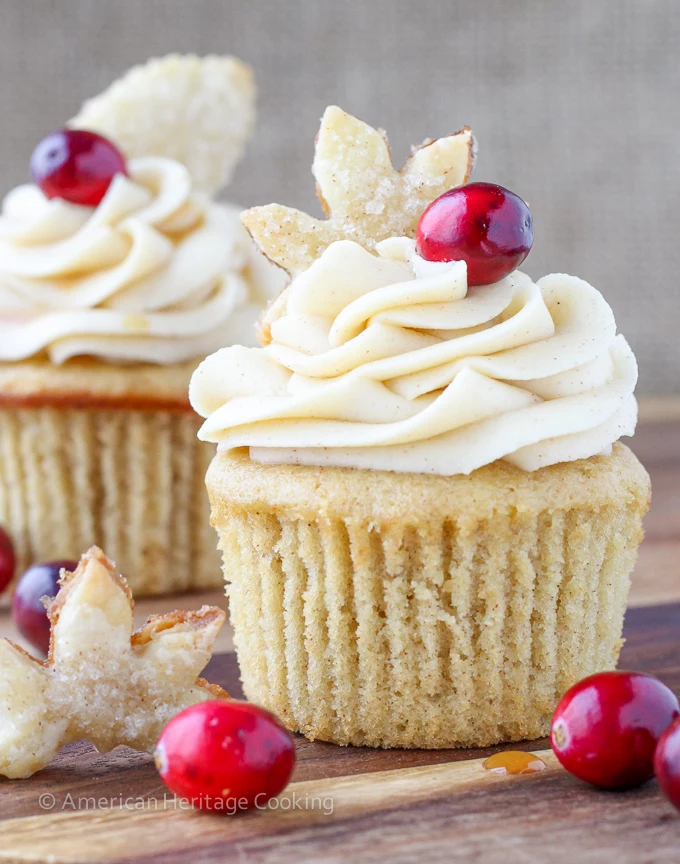two pale brown cupcakes, with pale yellow icing, with leaf-shaped cookies and cranberries on top, on wooden table with more cranberries