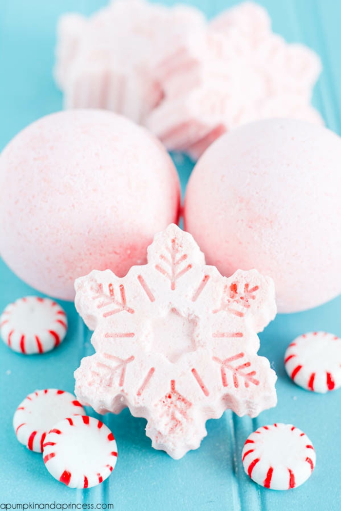 two round pale pink bath bombs, one snowflake shaped pale pink bath bomb, two more in background, five peppermint candies in foreground, all placed on blue surface