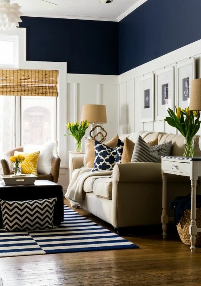 two tone living room walls, dark navy blue and white wall with plaster details, dark blue and white striped rug, cream colored sofa with yellow and blue cushions, brown chair with yellow and white cushions