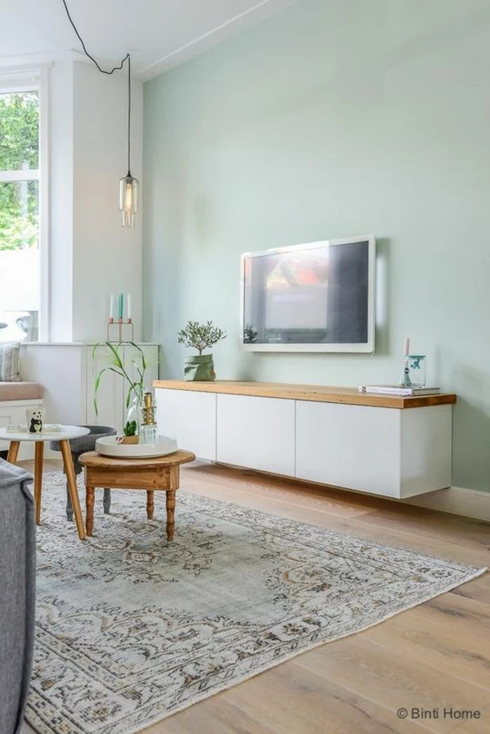 two tone living room walls, pale greenish-blue wall, opposing white wall with window, pale wooden floor and pale bluish carpet, two small coffee tables, white cupboard with wooden surface, lamp tv and plants 