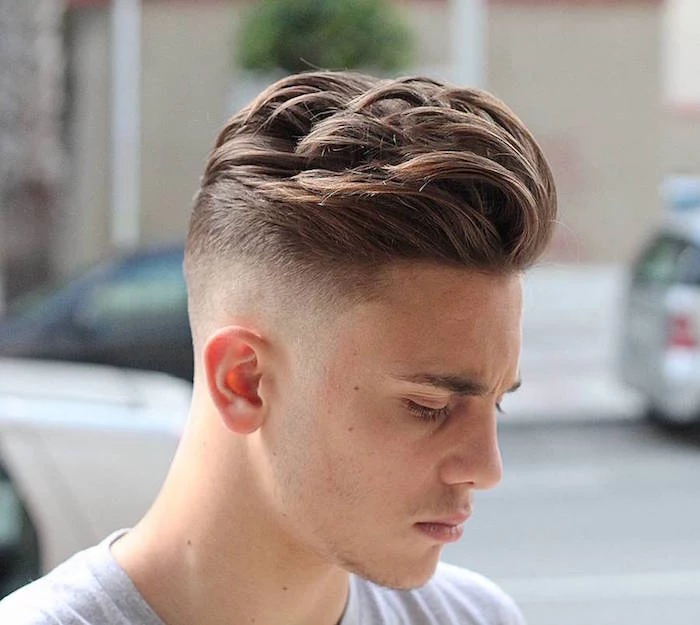 undercut fade, young man with chestnut hair, long and wavy on top and trimmed very short on the side, wearing light grey t-shirt facing sideways and looking serious