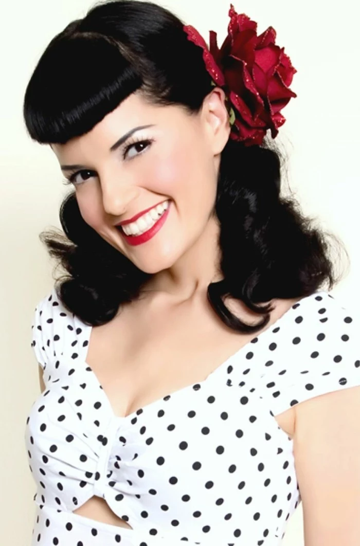 betty bangs, smiling woman with white teeth and bright red lipstick, shoulder-length wavy hair and a fake red rose ornament, white top with black polka dots