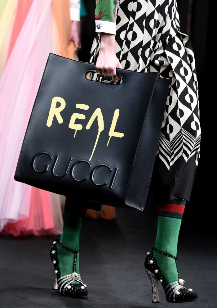 big black bag with inscription reading real gucci, held by woman in black and white dress with geometric pattern, green red and white sleeves, green and red socks and black and white shiny heels