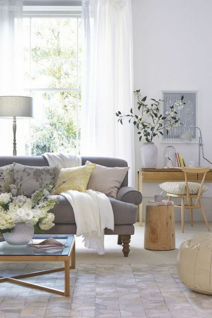 room with pale walls, grey sofa with four yellow and grey cushions and white blanket, wooden desk and chair, wooden coffee table with glass top, window with white curtains