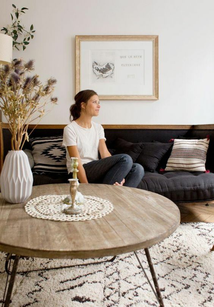 round wooden table with small crocheted cover, two candlesticks and a white vase with dry thistles, cream and dark brown carpet, woman sitting on dark blue sofa, white wall with framed image