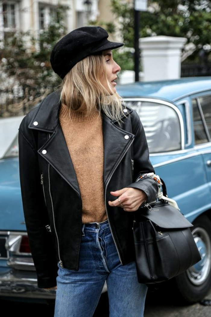 blonde woman with shoulder-length hair and black cap, wearing light brown sweater and acid wash blue jeans, with black leather biker jacket, holding a small black backpack