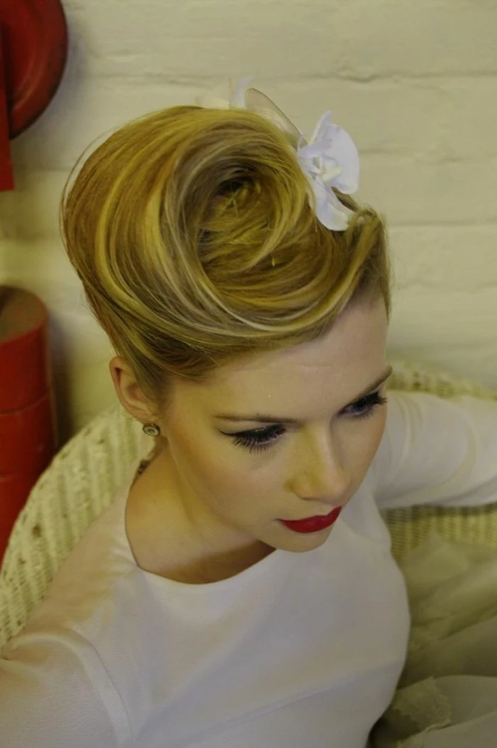 blonde woman with vintage swirly updo, and white hair ornament, wearing white top red lipstick and black eye make up, sitting on chair and seen from above