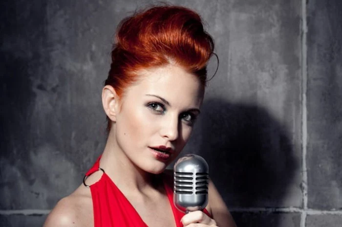 rockabilly hairstyles, red-haired woman with hair done up, dark red lipstick blush and eye makeup, red dress with metal detail, holding vintage microphone