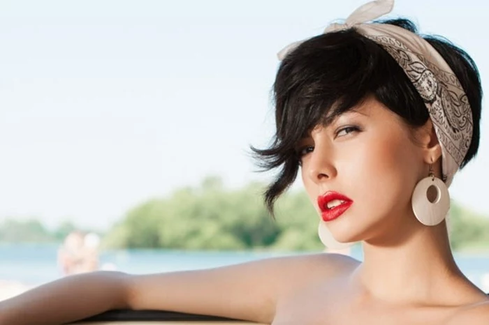 rockabilly hairstyles, serious-looking woman with long black bangs, hair tied with white and black bandanna, big white earrings and bright red lipstick, water and trees in background