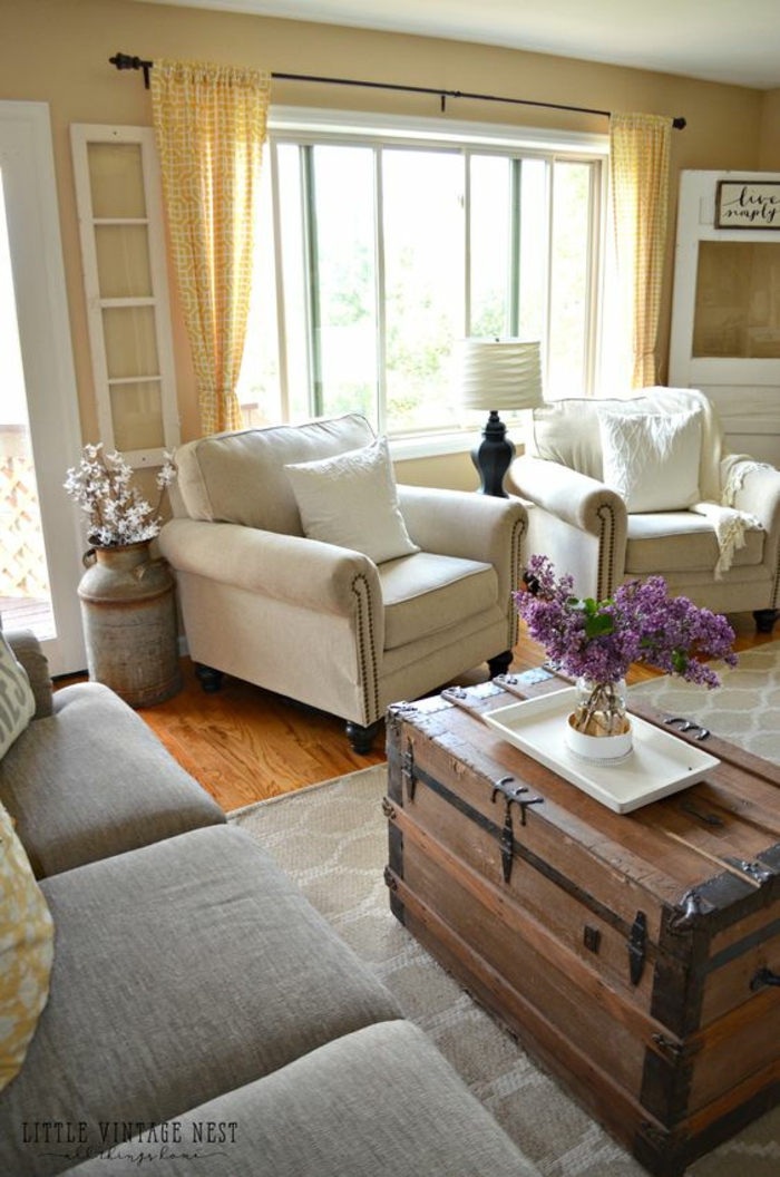 living room paint colors, pale yellow walls and curtains, light grey sofa and two cream colored chairs with pillows, big wooden chest used as table, white tray and a vase with lilacs