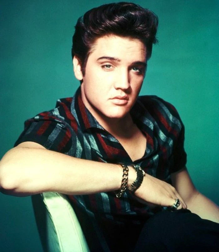 colorized photo of elvis presley leaning on white chair, dark gelled up hair, red grey and black striped short sleeved shirt, chain bracelet ring and wrist watch