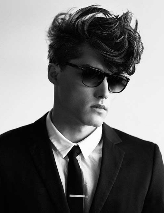 black and white image of young man wearing dark sunglasses, gelled up messy hair, dark blazer over white shirt, black necktie with light pin