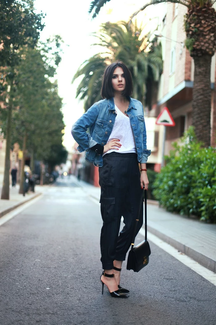 young woman with dark cropped hair, wearing denim jacket over white top, black cargo pants and strappy high heels, hand on hip and small black bag