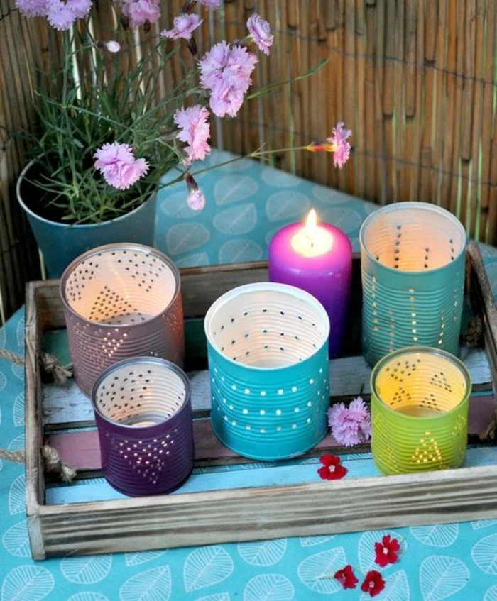 five cans, painted in different colors, with small holes forming shapes, placed on a wooden tray, near a lit purple candle