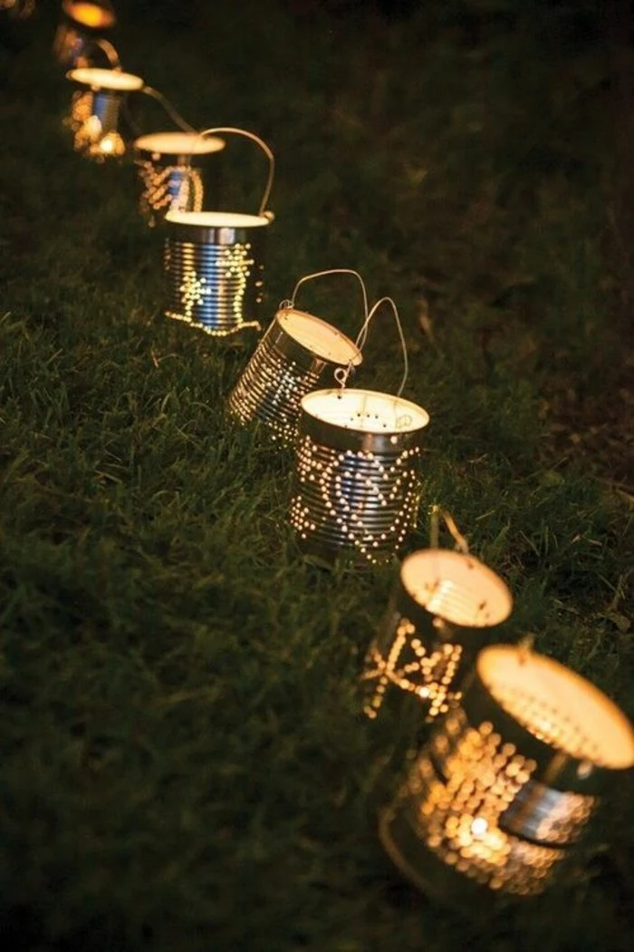 many luminaries made from plain tin cans with holes, with lit candles inside and with wire handles, placed on grassy area