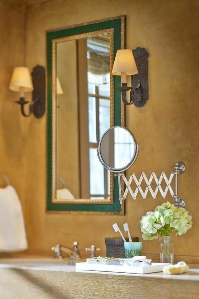 bathroom makeovers, sienna yellow wall, mirror in green frame, two wall lights, magnifying mirror near toothbrushes and flowers