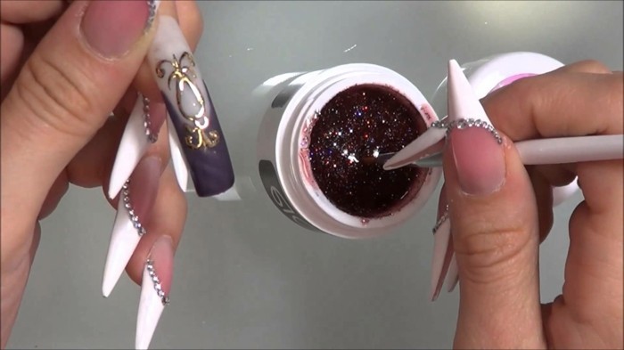 nail designs with rhinestones, two hands with long nails, with rhinestones and sharp white tips, holding fake nail and paint brush dipped in paint