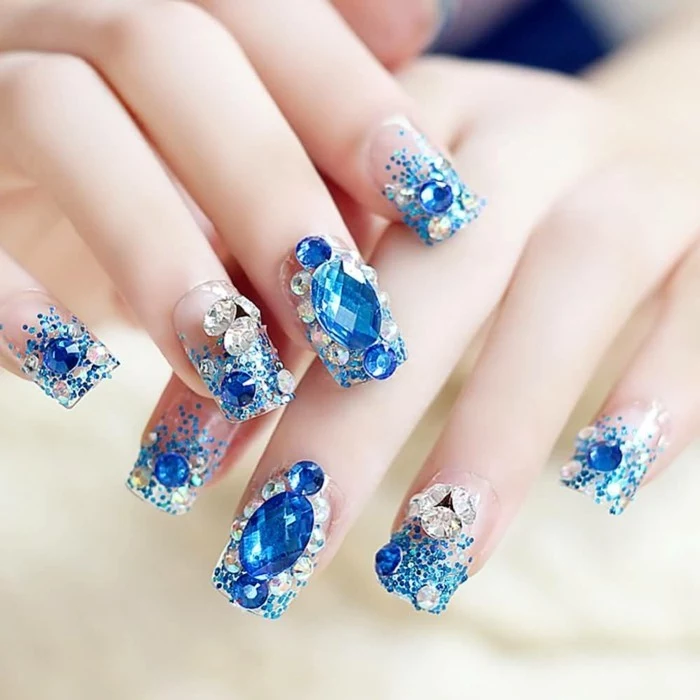 rhinestone nail art, close up on two hands, decorated with fake nails featuring big blue gems, white rhinestones and blue glitter