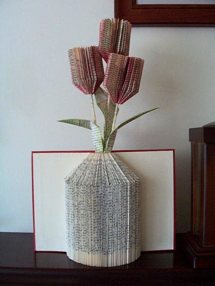 book folding art, book with red and white hard covers, opened to reveal a vase shape, made from folded pages, with paper flowers, made from more cut and folded pages, painted red and green