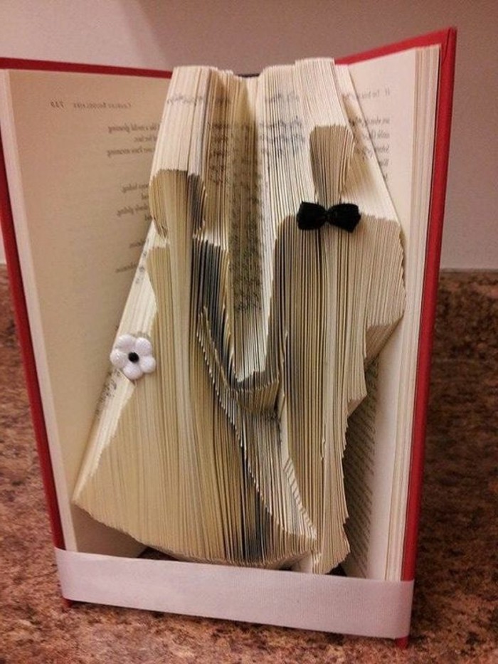 bookfolding, bride and groom holding hands, made from folded pages, decorated with small white flower and tiny black bowtie, inside an open book with hard red covers, tied with white ribbon